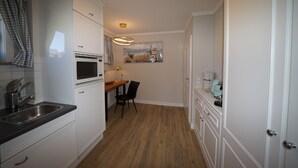 A compact kitchen with white cabinets, a wooden countertop, a microwave, a sink, a dining table with chairs, and a painting on the wall. - Strandvilla Wohnung Strand-loft (Sylt)