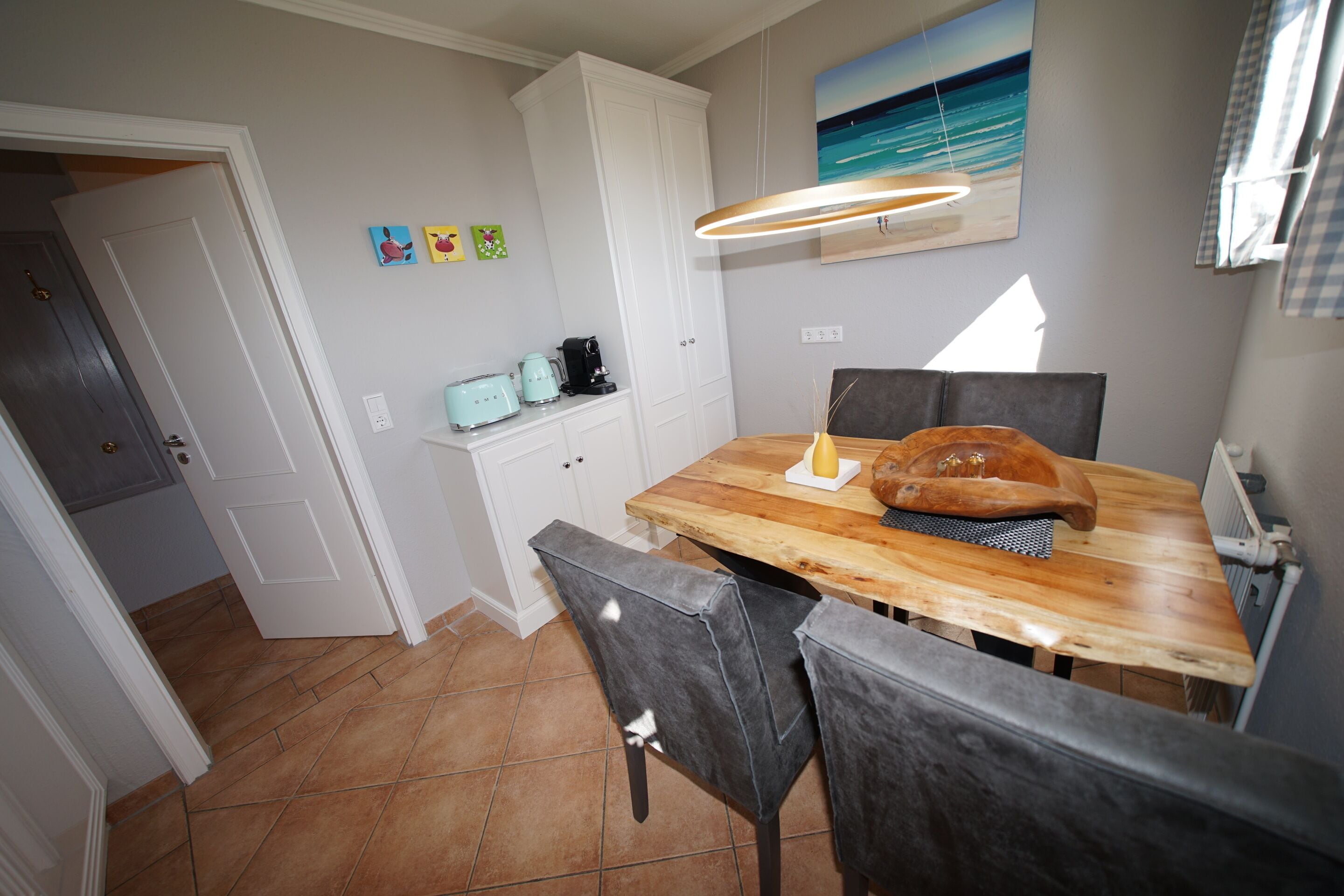 A dining area with a wooden table, two chairs, a painting of a beach, and a ceiling light fixture.