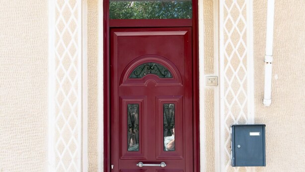 Una puerta roja con el número "55" encima, un buzón a la derecha y un pequeño escalón que lleva a la puerta.