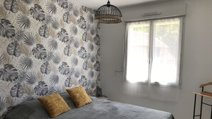 Une chambre à coucher avec un lit, deux oreillers, une table de chevet, une lampe, une fenêtre avec des rideaux et un luminaire suspendu.