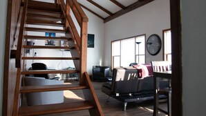 Een ruime woonkamer met een houten trap, een zithoek, een eettafel en een groot raam met een klok.