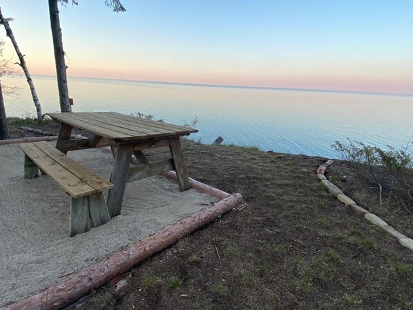 Une table de pique-nique en bois, avec des bancs, installée sur une zone sablonneuse, offrant une vue sur l’eau.