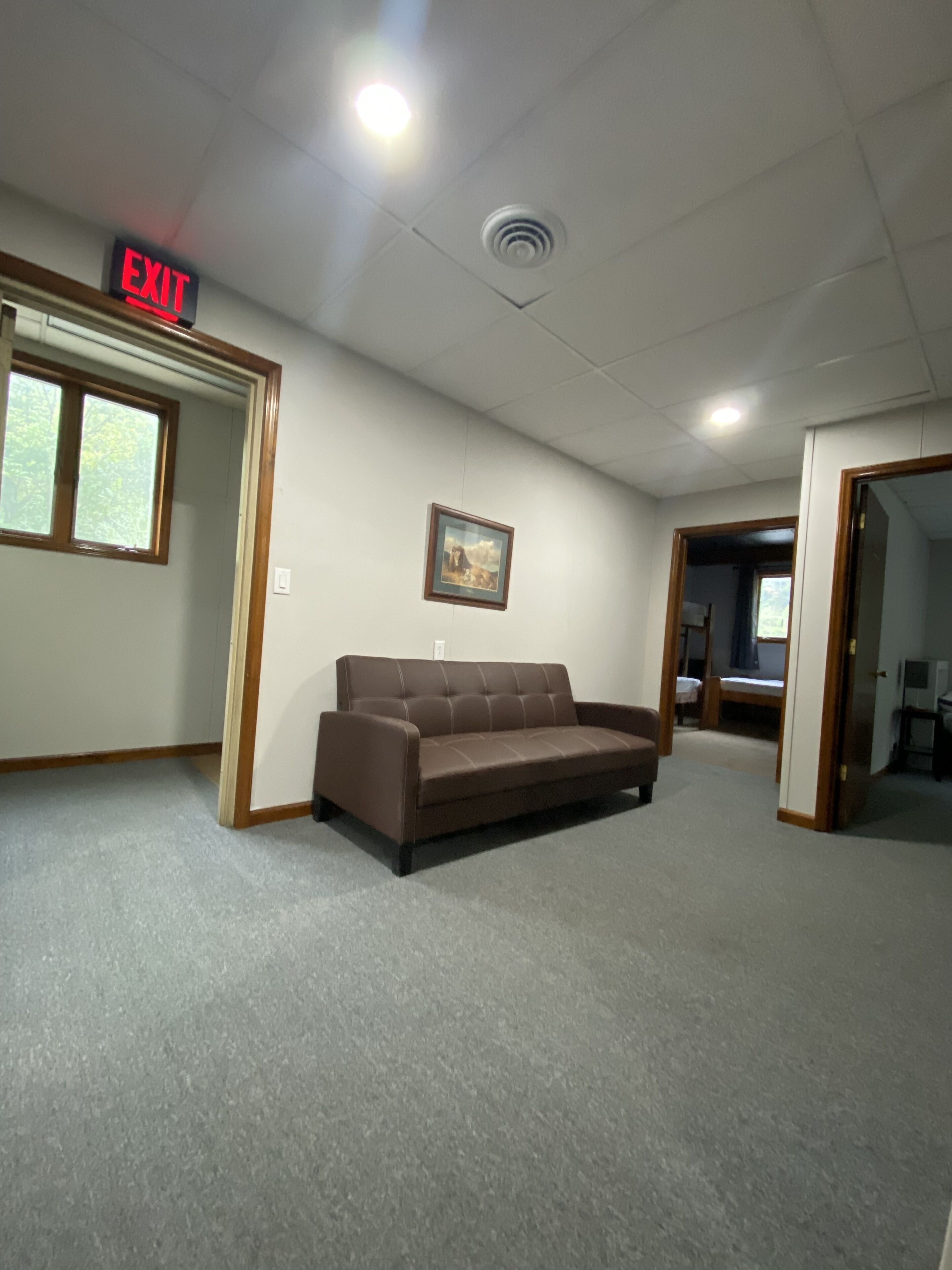 A room with a brown sofa, an exit sign, a framed picture on the wall, and a window.