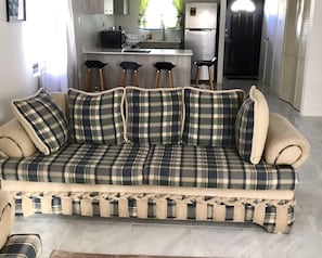 A sofa with a plaid pattern, a kitchen area with a refrigerator and bar stools, and a door in the background.