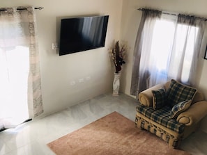 A living room with a flat-screen TV mounted on the wall, a beige sofa with a plaid throw, a brown rug, and a white vase with dried flowers.