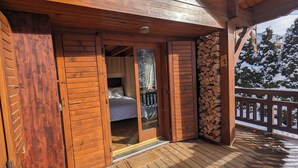 A wooden cabin with a balcony, a bed with a white comforter, and a pile of firewood. - Authentic chalet in the heart of Serre Chevalier, 300m from the slopes (Saint-Chaffrey)