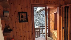 A wooden cabin interior with a wooden table, a chair, a bed, and a sliding glass door leading to a balcony with a wooden railing. - Authentic chalet in the heart of Serre Chevalier, 300m from the slopes (Saint-Chaffrey)