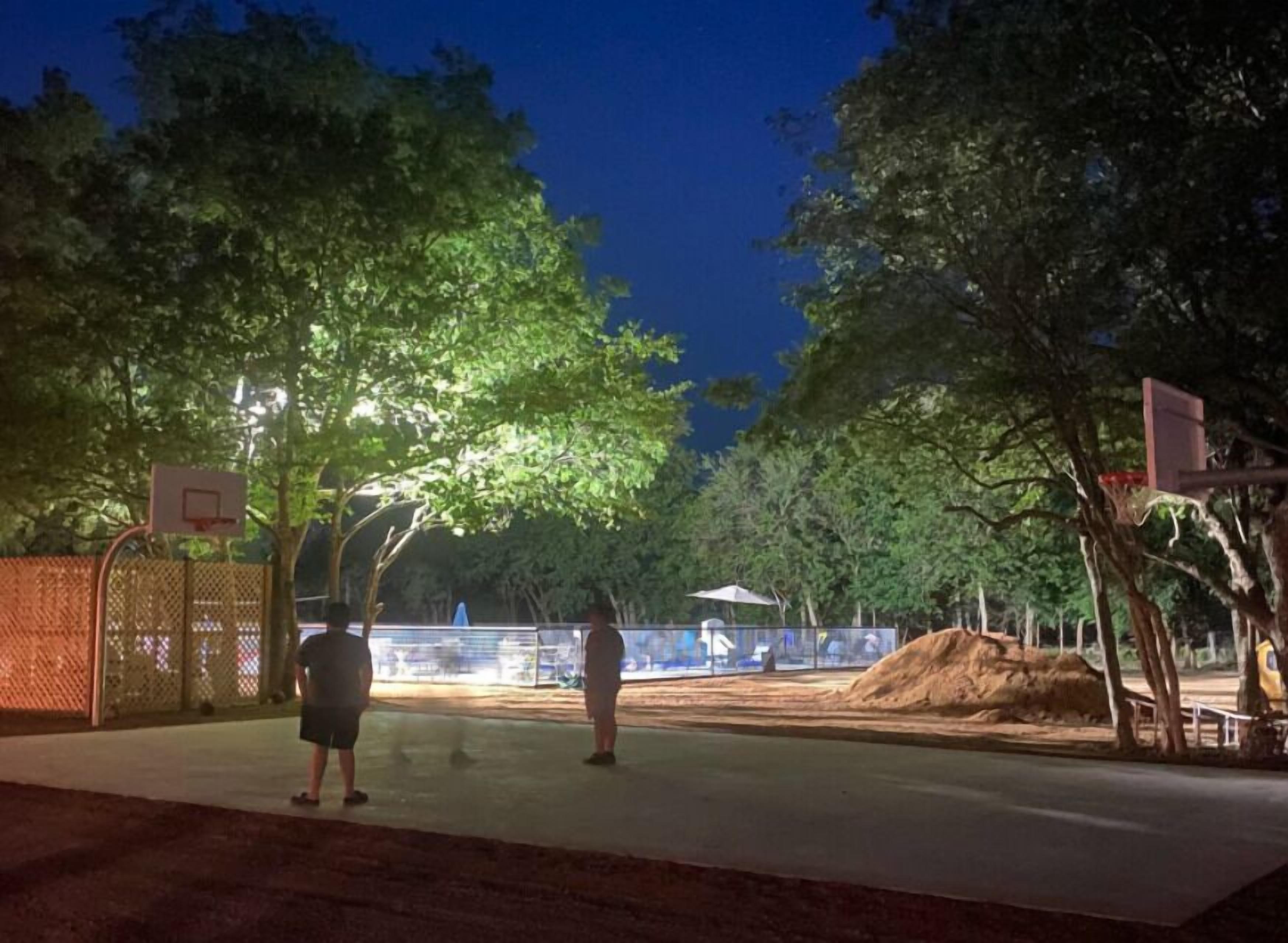 Un terrain de basket en plein air, illuminé la nuit, avec des arbres et une clôture en arrière-plan.