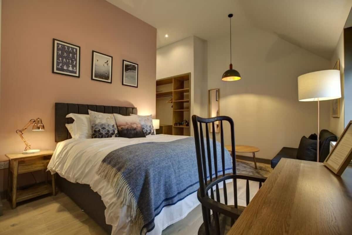 A bedroom with a bed, bedside tables, a chair, a desk, a lamp, a wardrobe, and a hanging light fixture.