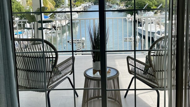 Un balcon avec deux fauteuils en osier et une petite table, donnant sur une marina où sont amarrés des bateaux, et sous un ciel dégagé.