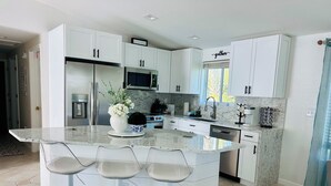 Une cuisine moderne avec des meubles blancs, un grand îlot central doté de tabourets de bar, des appareils en acier inoxydable et une fenêtre ornée d’une inscription indiquant « lieu de rassemblement ».