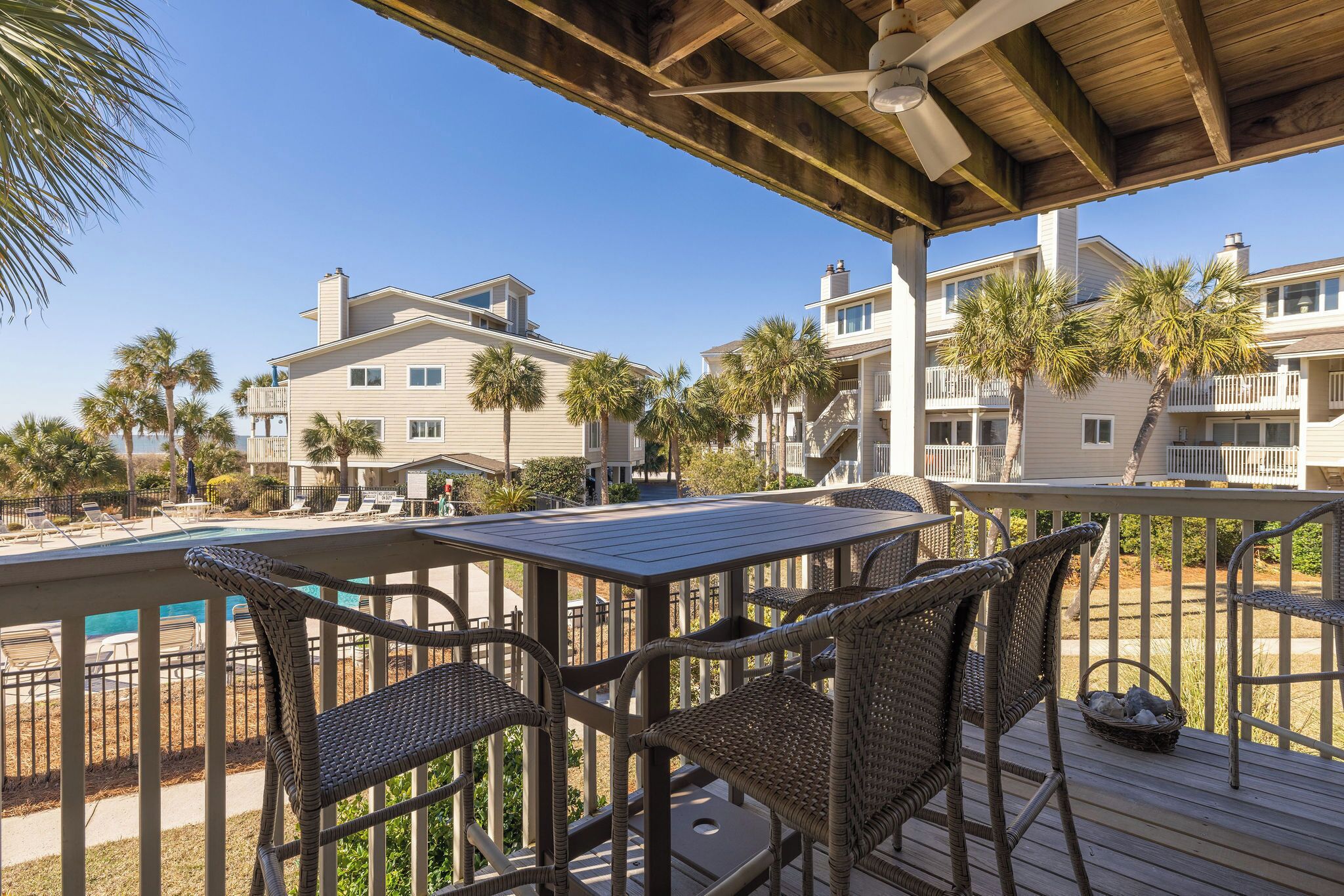 A wooden deck with wicker furniture, overlooking a pool and beachfront buildings.