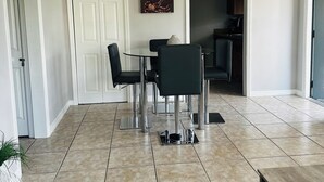 A dining area with a table and chairs, a painting on the wall, and a kitchen in the background. - Spacious and Bright 2bed, washer/dryer, back yard (Delray Beach)
