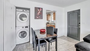 A compact living space with a washing machine, a dining table with chairs, a red bowl, and a door leading to a kitchen area. - Spacious and Bright 2bed, washer/dryer, back yard (Delray Beach)
