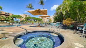 A swimming pool with a mosaic design, surrounded by lounge chairs and umbrellas, with palm trees and buildings in the background.