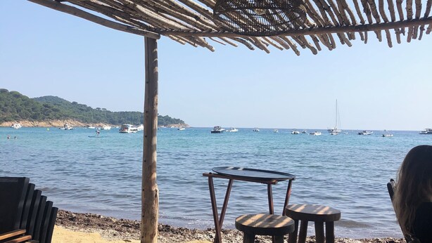 Un área de estar junto a la playa con una pérgola de madera, una mesa y dos taburetes.