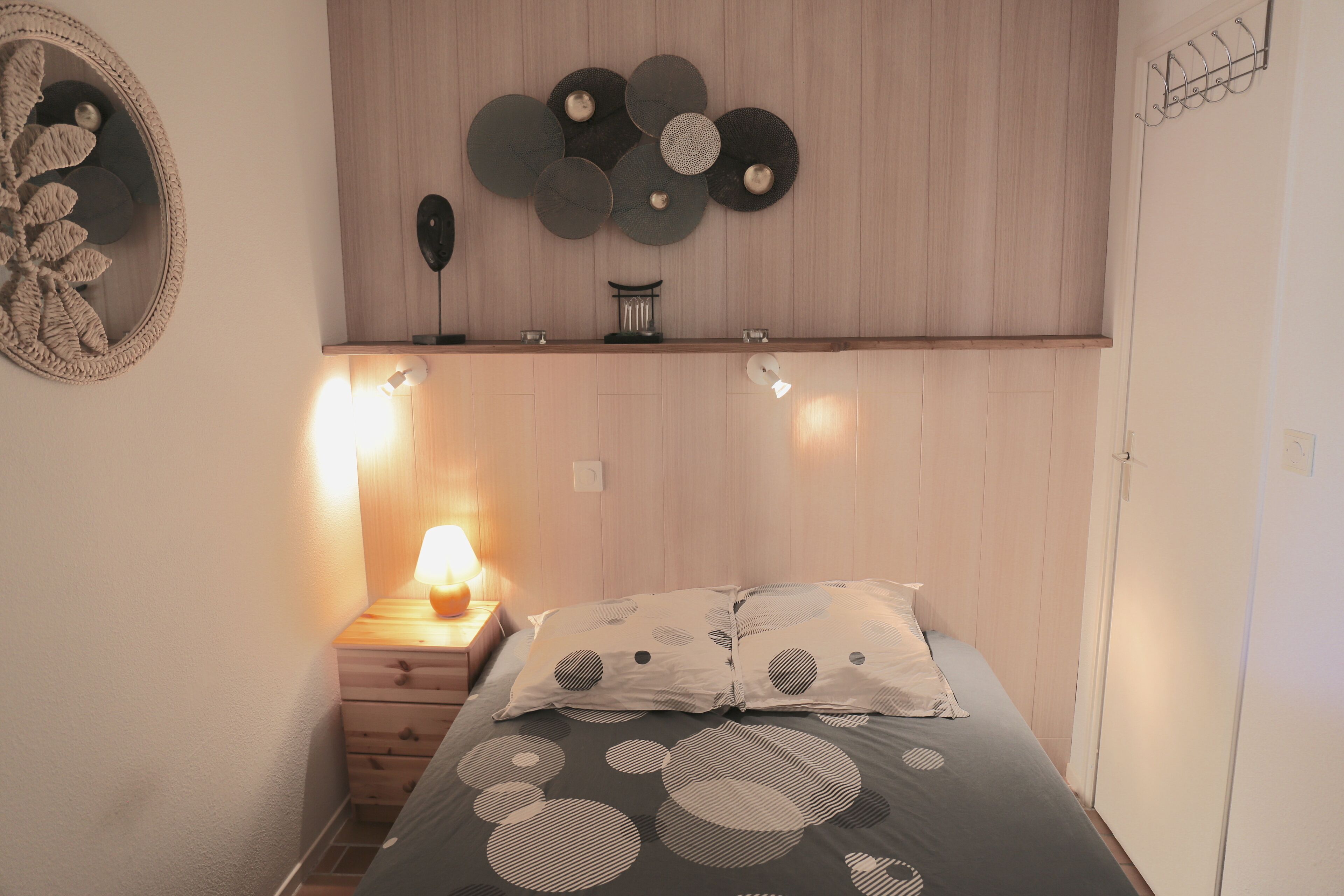 A bedroom with a bed, a nightstand with a lamp, a wall-mounted shelf with decorative items, and a wooden headboard.