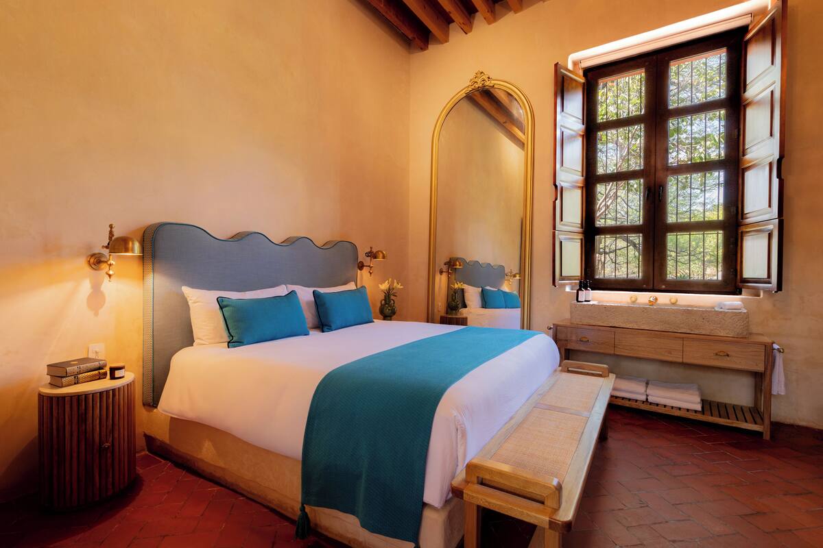 A neatly made bed with a blue and white bedspread, a wooden bench, a bedside table with a lamp, and a large mirror above a window.