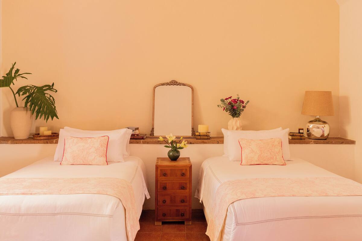 A hotel room with two beds, a nightstand, a mirror, a vase with flowers, and a lamp.