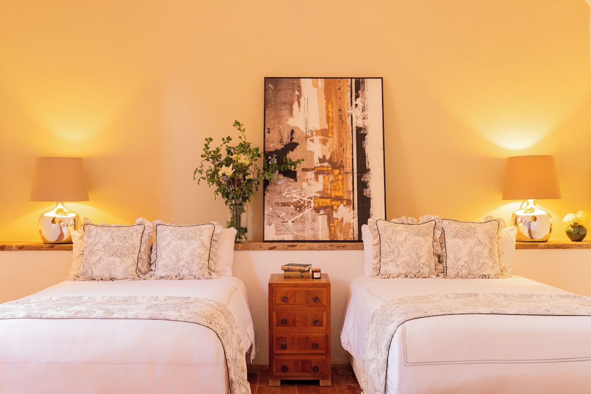 A hotel room with two beds, a wooden nightstand, a painting on the wall, and two lamps.