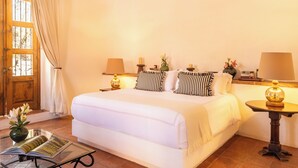 Un dormitorio con una cama grande, dos mesitas de noche con lámparas, una mesita con un jarrón y una ventana con cortinas.