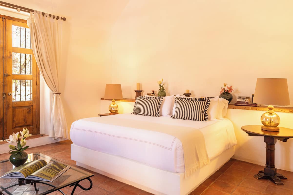 A bedroom with a large bed, two bedside tables with lamps, a small table with a vase, and a window with curtains.