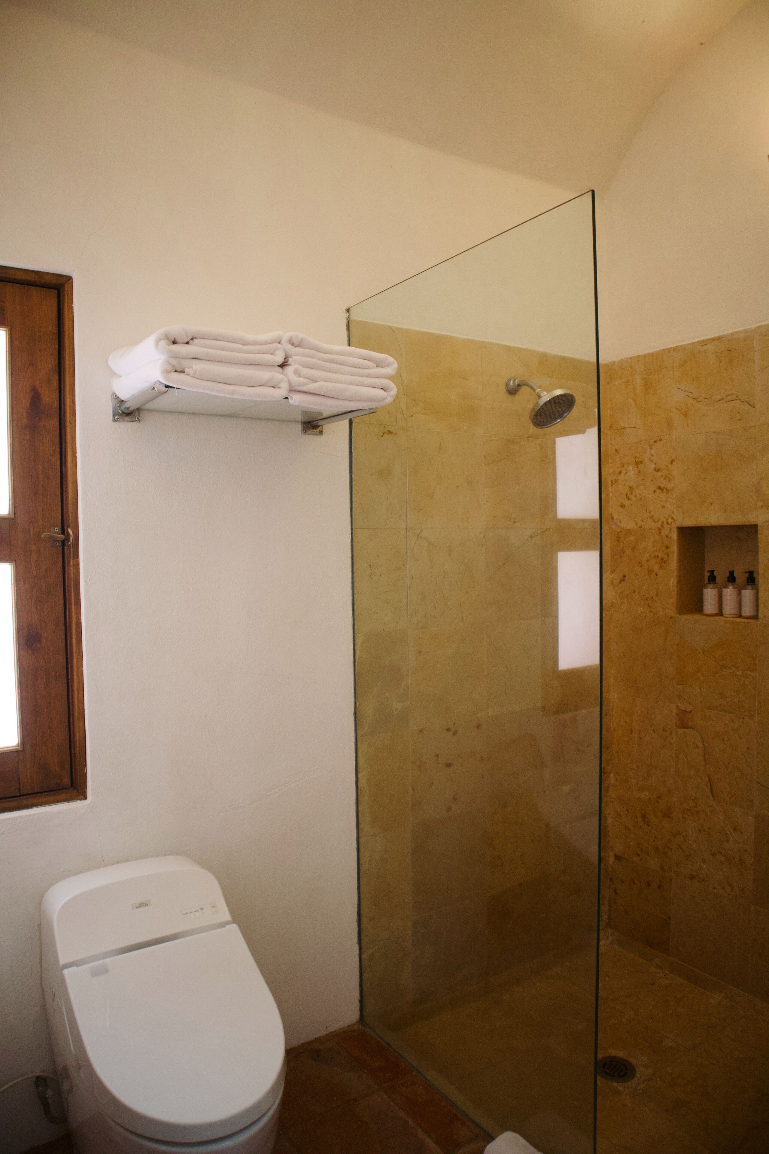 A bathroom with a glass-enclosed shower, a toilet, and a shelf with folded towels.