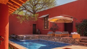 Una piscina con bordo in pietra, una pergola in legno, un edificio rosso e area salotto all'aperto con un tettoia.