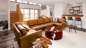 A spacious living area with a large L-shaped sectional sofa, a round coffee table, and a wooden accent piece. A staircase leads to an upper level, and a kitchen area is visible in the background. - A 160 Acre Luxury Modern Ranch (Belgrade)