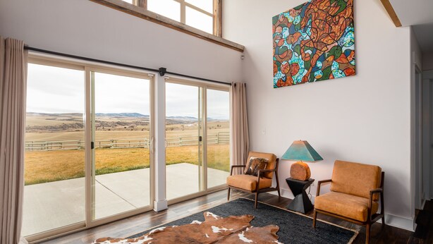 A spacious room with a large sliding glass door leading to a scenic outdoor view, a cowhide rug, two leather chairs, a table with a lamp, and a colorful abstract painting on the wall. - A 160 Acre Luxury Modern Ranch (Belgrade)