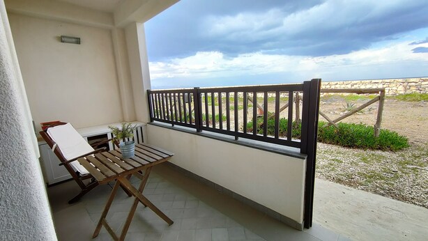 A balcony with a wooden table and chairs, a potted plant, and a view of a garden and sky. - Accommodation Mediterraneo Verde - Marzamemi (Marzamemi)