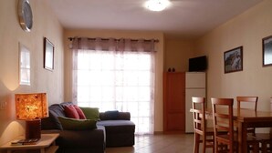 En stue med sofa, et lille bord med lampe, spisebord med stole, fjernsyn på et skab og et vindue med gardiner.