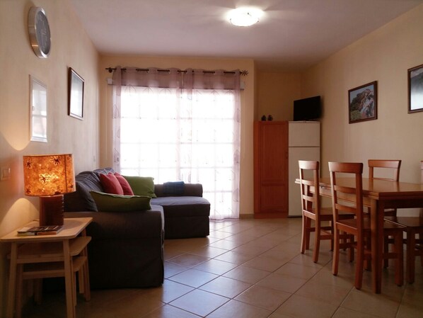 A living room with a sofa, a small table with a lamp, a dining table with chairs, a TV on a cabinet, and a window with curtains.
