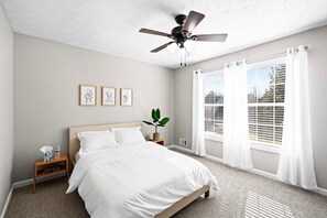 A bedroom with a bed, a nightstand, a ceiling fan, a window with curtains, and a plant.