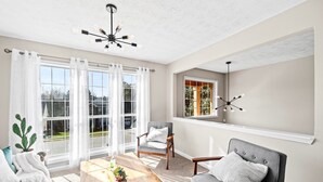 Un salon avec une table basse en bois, deux fauteuils gris, un canapé blanc, un ventilateur de plafond et une vue sur l’extérieur grâce à de grandes fenêtres.