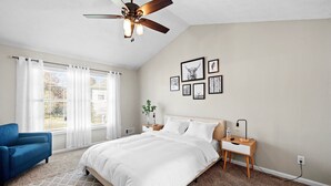Une chambre à coucher avec un lit, une chaise bleue, une table de chevet, un ventilateur de plafond fixé au mur et des tableaux encadrés.
