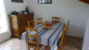 Een eetgedeelte met een tafel gedekt voor vier personen, omringd door houten stoelen. Op de tafel staat een mand met fruit en erboven hangen twee ingelijste foto's aan de muur.
