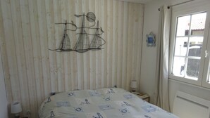 Een slaapkamer met een bed, een nachtkastje, een lamp, een raam met gordijnen en een wanddecoratie van zeilboten.