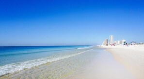 A beach with clear blue water, sandy shore, and buildings in the background.