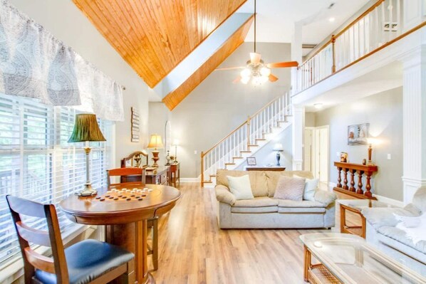 Relax in a spacious living area with a high ceiling and exposed beams, featuring a sofa and coffee table, staircase, and large windows with blinds.