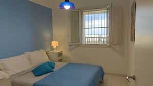 Een slaapkamer met een bed, nachtkastje, lamp, raam met uitzicht op het strand en een deur.