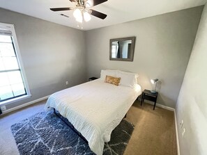 Een slaapkamer met een bed, een plafondventilator, een nachtkastje met lamp, een raam en een spiegel aan de muur.