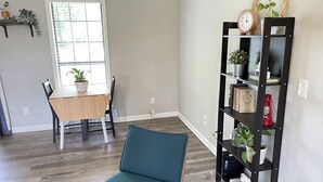 A room with a dining table, chairs, a teal sofa, a bookshelf, and a window with blinds. - Spacious 4 bedroom home near Charleston & beaches! (Ladson)