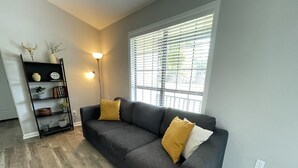 A living room with a dark grey sofa, two yellow cushions, a black metal shelf with decorative items, a floor lamp, and a window with blinds. - Spacious 4 bedroom home near Charleston & beaches! (Ladson)