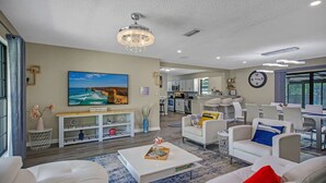A modern living room with a white sofa, a coffee table, and a large painting of a coastal scene. A kitchen is visible in the background with white cabinets and a dining area with a table and chairs. - SWEET HOME With Private Pool, Patio,& Huge backyard. (Orange Park)