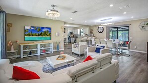 A modern living room with a white sofa, a coffee table, and a TV mounted on the wall. There is a dining area with a table and chairs, and a kitchen in the background. - SWEET HOME With Private Pool, Patio,& Huge backyard. (Orange Park)