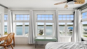 A room with a bed, a desk, and a chair. There are three windows with white curtains, and a view of a body of water and dock. - Lakeside, New Large Dock & Mooring Ball. On beautiful  Lake Charlevoix 🥂 (East Jordan)
