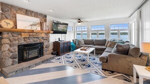 A living room with a stone fireplace, a large sofa, and a view of the water. - Lakeside, New Large Dock & Mooring Ball. On beautiful  Lake Charlevoix 🥂 (East Jordan)