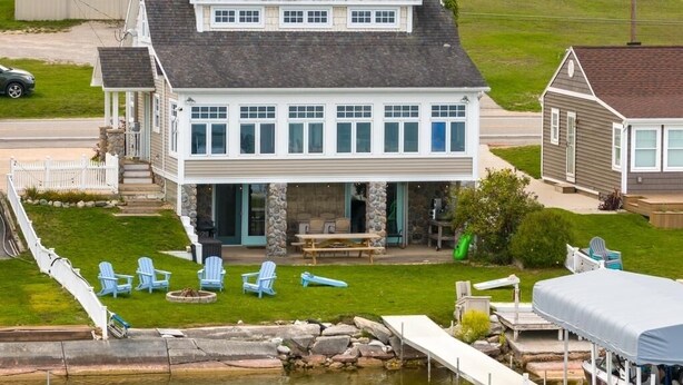 A two-story house with a dock extending into a pond, surrounded by a well-maintained lawn and outdoor seating area. - Lakeside, New Large Dock & Mooring Ball. On beautiful  Lake Charlevoix 🥂 (East Jordan)