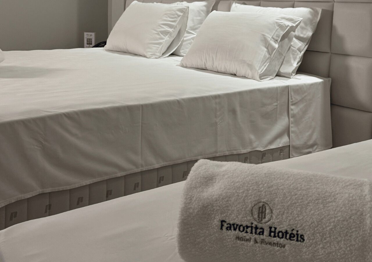 Dos camas con sábanas y almohadas blancas, un cabecero y una toalla con el texto 'Favorita Hotéis'.